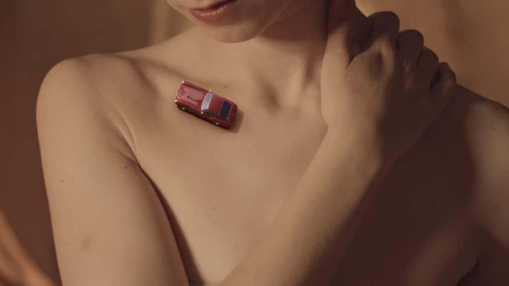 Ein Videostill aus dem Musikvideo "Ella". Ein kleines, rotes, animiertes Auto fährt über das Schlüsselbein einer nackten Frau