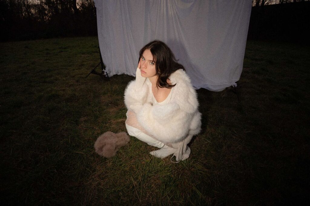 Ein Bild der Sängerin Diemarcha auf einer Wiese vor einer weißen Leinwand.