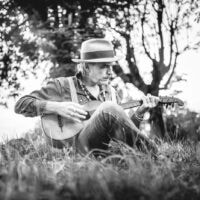 Schwarzweiß Porträt vom Musiker Ernst Molden mit Akustikgitarre auf Wiese vor Bäumen sitzend