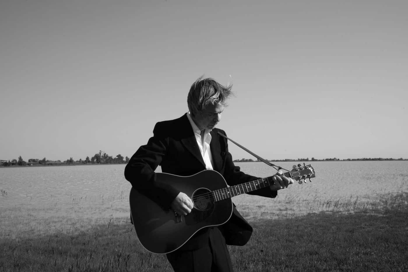 Ein schwarz-weißes Bild des Musikers Hannes Duscher mit einer Akkustikgitarre auf einem Feld stehend