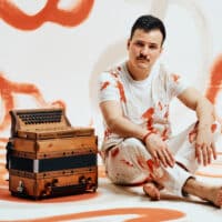 Der Musiker Jakob Steinkellner mit seiner Steirischen Harmonika auf orange-weißem Hintergrund