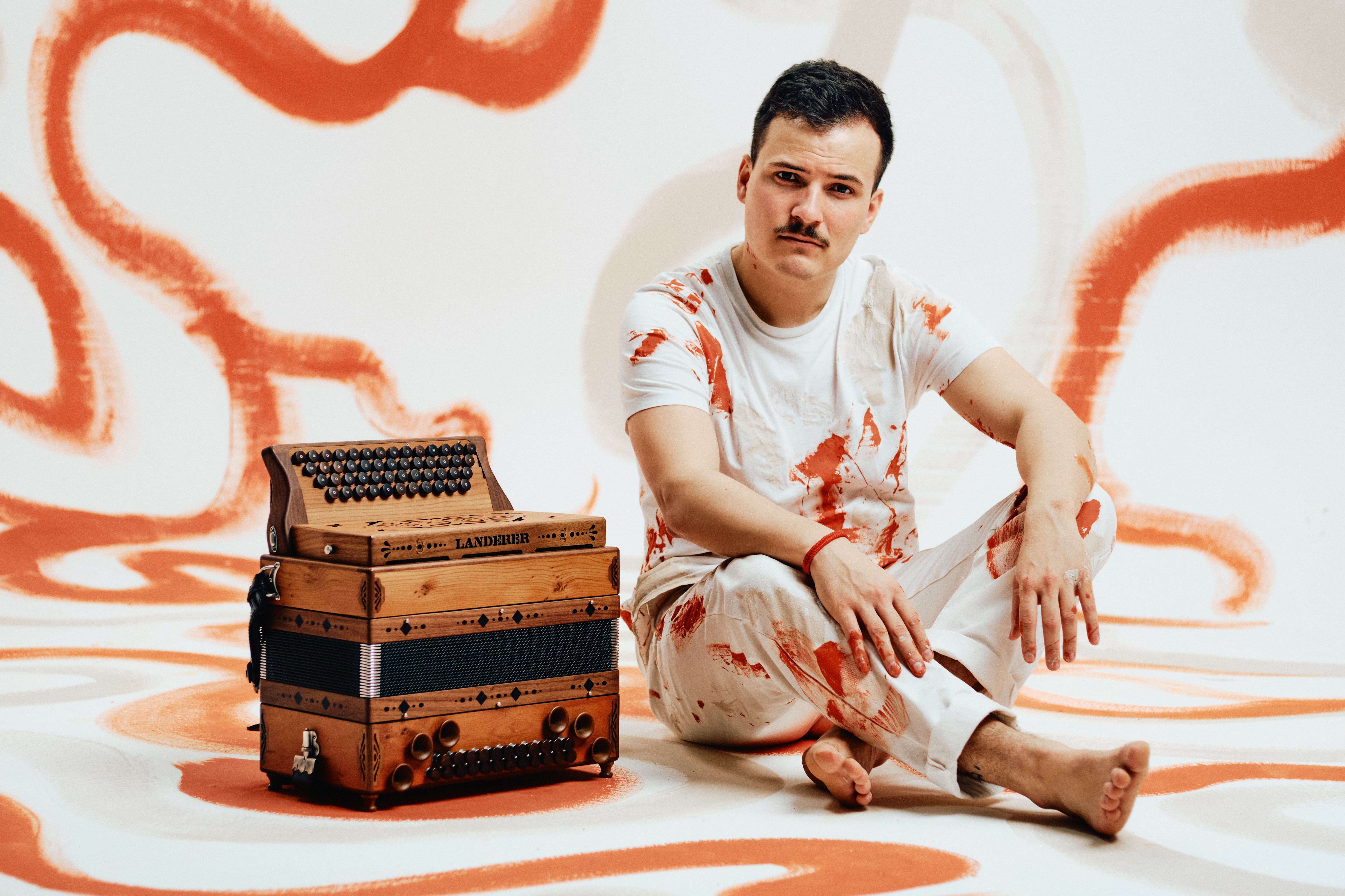 Der Musiker Jakob Steinkellner mit seiner Steirischen Harmonika auf orange-weißem Hintergrund