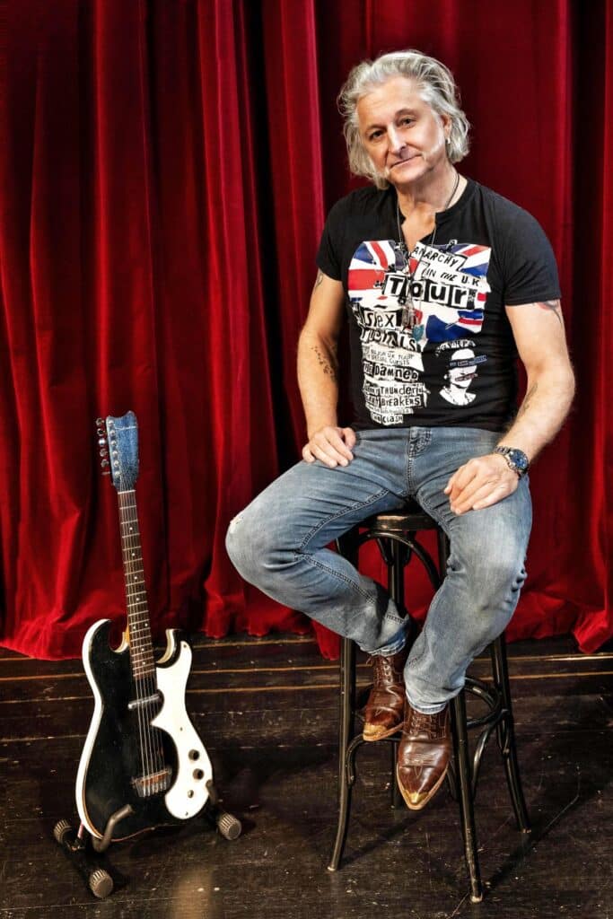 Bild Sir Oliver Mally auf einem Barstuhl auf der Bühne vor rotem Vorhang sitzend mit E-Gitarre neben ihm platziert