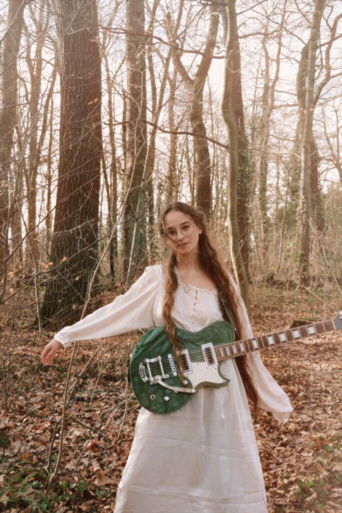 Bild der Liedermacherin Sodl in weißem Kleid im Wald mit Gitarre stehend