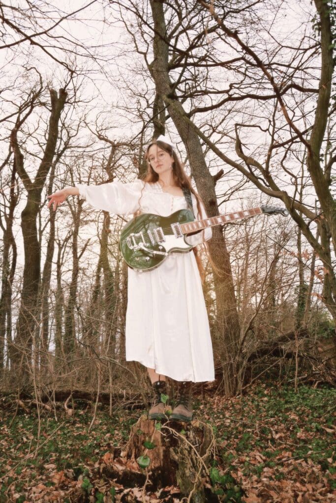 Bild der Liedermacherin Sodl in weißem Kleid im Wald mit Gitarre stehend