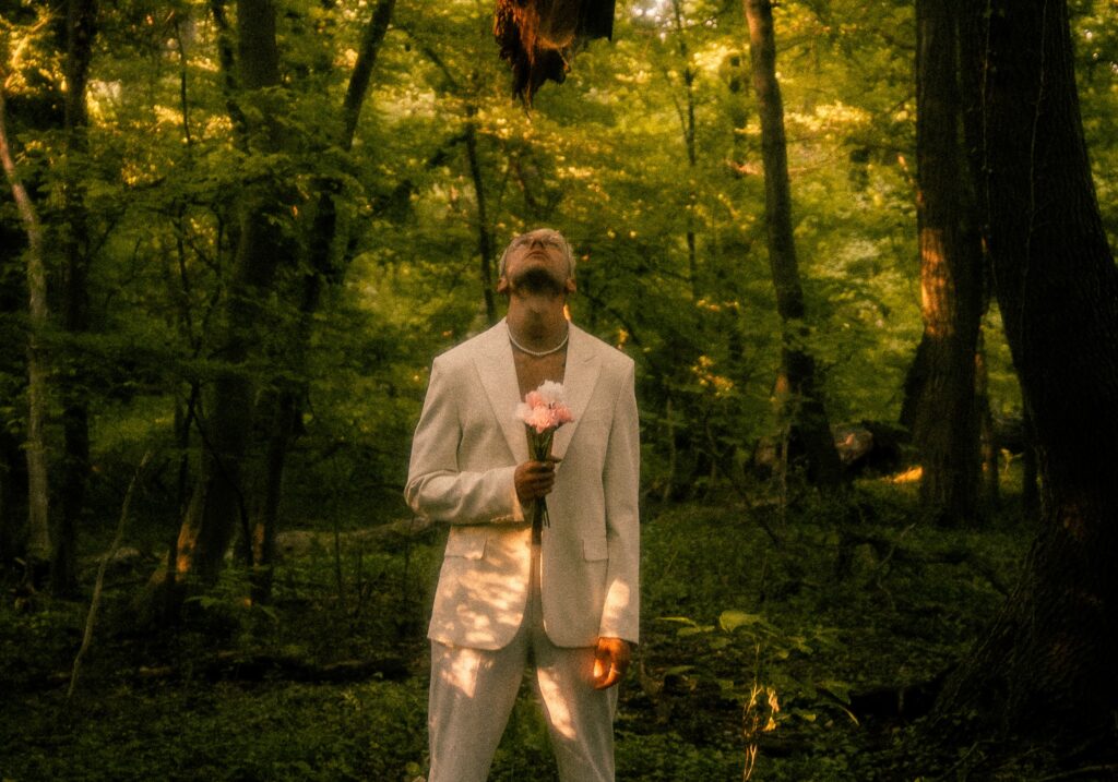 Der Künstler palffi in einem weißen Anzug steht im Wald mit einem Blumenstrauß unter einem abgebrochenen Ast.